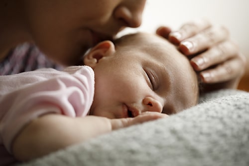 A mother kissing the back her sleeping baby's head with her fingertips resting gently on the top of the baby's head.